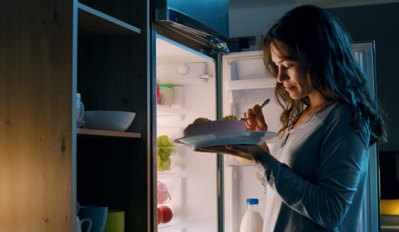 O femeie care mănâncă dintr-o farfurie, în fața frigiderului