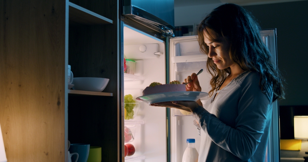 O femeie care mănâncă dintr-o farfurie, în fața frigiderului
