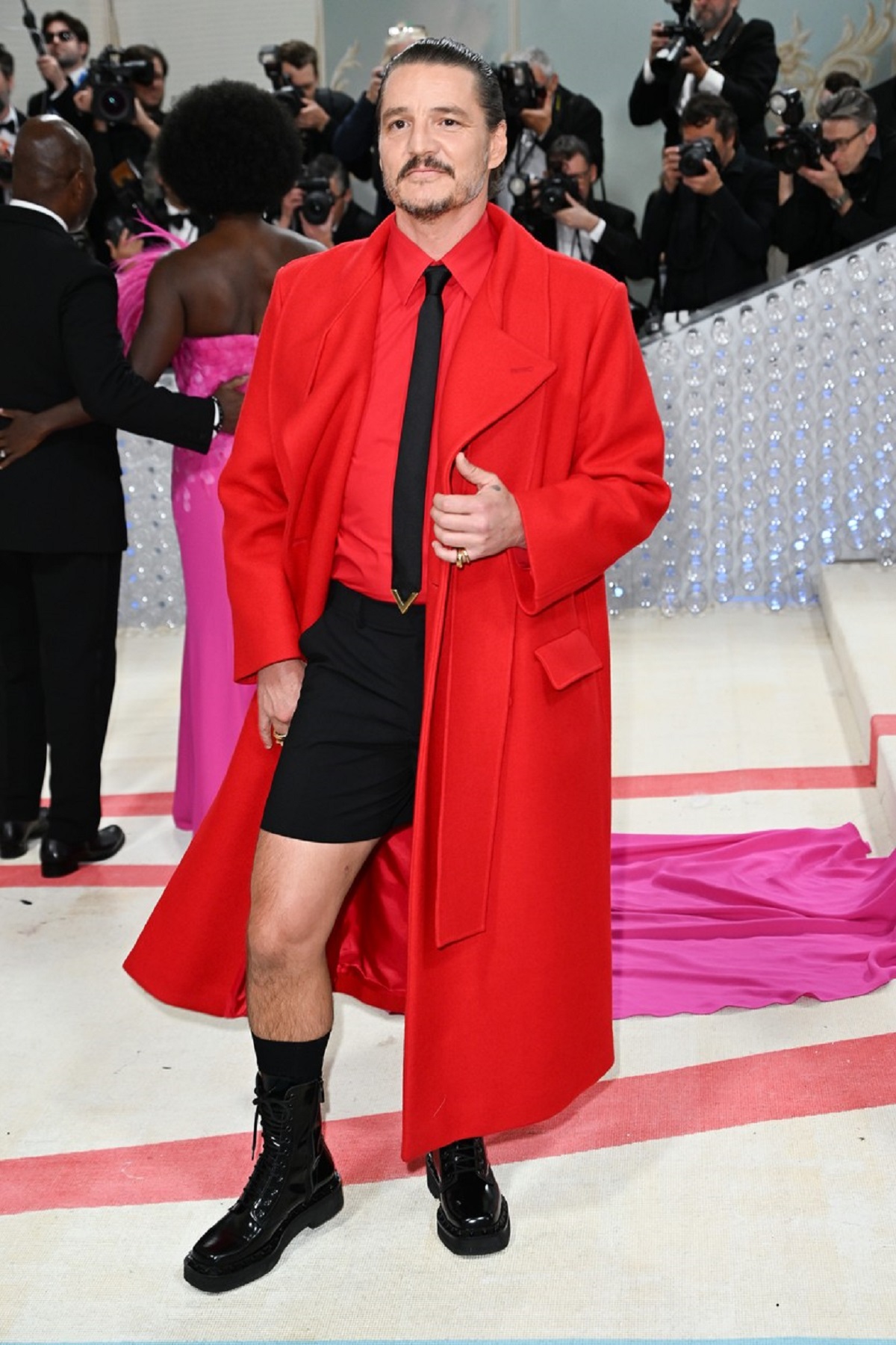 Pedro Pascal într-o pereche de pantaloni negri și un palton roșu pe covorul roșu de la Met Gala 2023