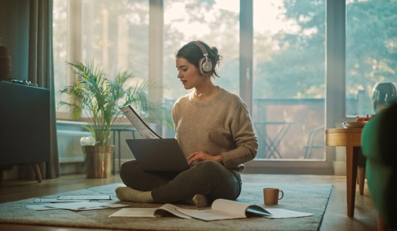 O femeie tânără stă pe podea, într-o cameră cu ferestre și o plantă, cu un laptop pe genunchi, căști pe urechi, iar în jur sunt reviste și o cană.