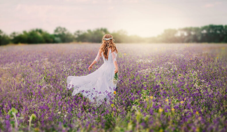 Fată frumoasă cu părul blond îmbrăcată într-o rochie lungă, albă stă într-un câmp cu fața la soare