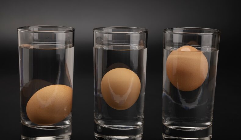 Trei pahare cu apă, fiecare cu un ou care plutește, fotografiate pe un fundal negru