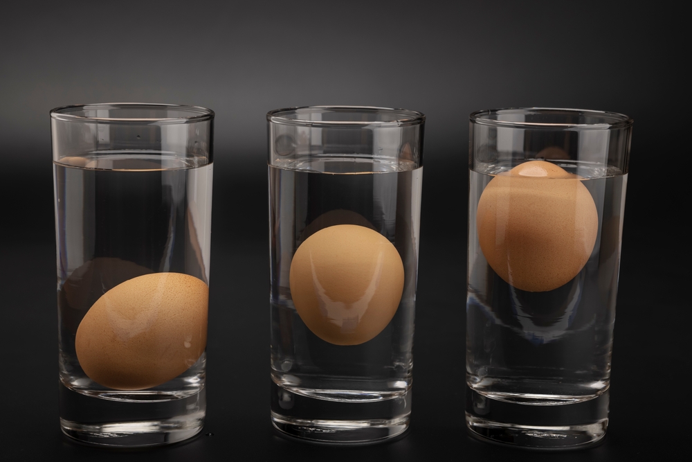 Trei pahare cu apă, fiecare cu un ou care plutește, fotografiate pe un fundal negru