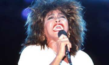 Tina Turner, pe scenă, fotografiată în timp ce cântă, într-un sacou alb