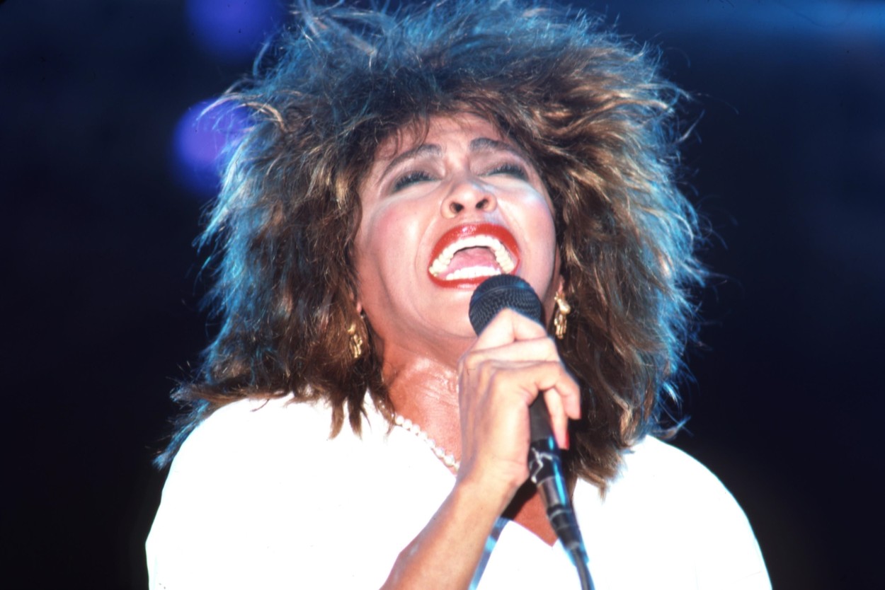 Tina Turner, pe scenă, fotografiată în timp ce cântă, într-un sacou alb