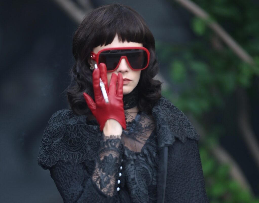 Cara Delevingne, cu ochelari de soare cu rame roșii, într-o ținută de culoare neagră