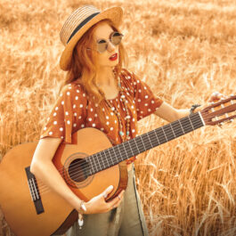 Fată frumoasă cu părul lung desprins, cu pălărie pe cap stă într-un lan de grâu cu o chitară în mână