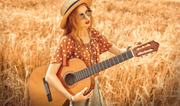 Fată frumoasă cu părul lung desprins, cu pălărie pe cap stă într-un lan de grâu cu o chitară în mână