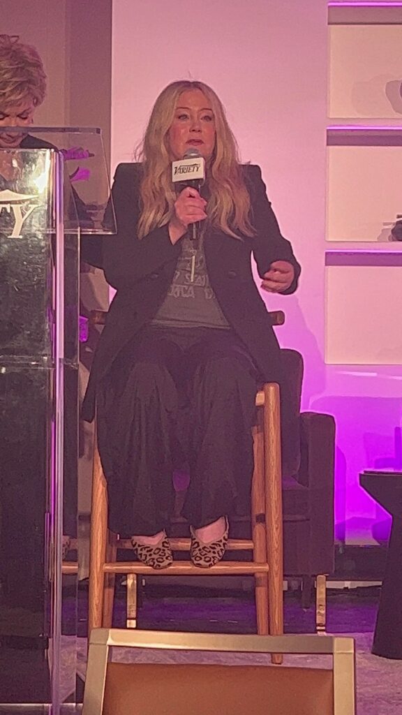 Christina Applegate, pe un scaun, îmbrăcată în negru, fotografiată în timp ce vorbește la un microfon