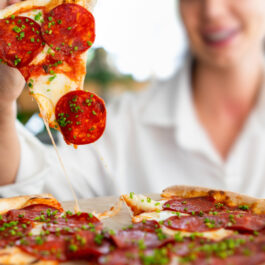 O femeie zâmbitoare rupe o felie de pizza cu salam, brânză și mirodenii