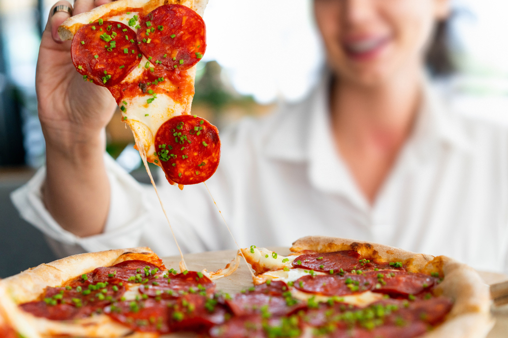 O femeie zâmbitoare rupe o felie de pizza cu salam, brânză și mirodenii