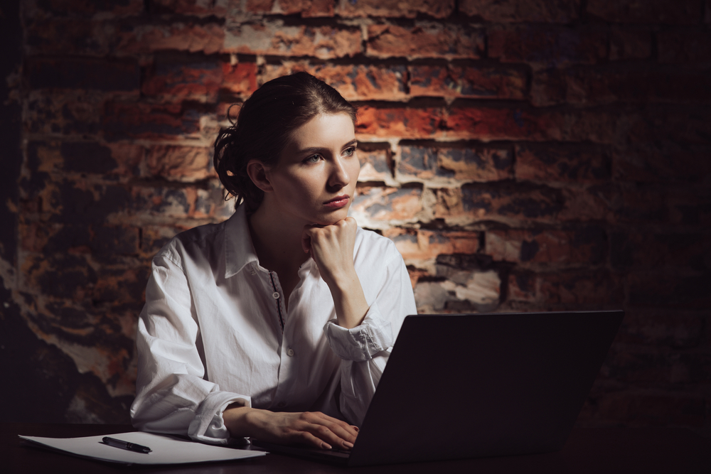 Fată îmbrăcată într-o cămașă stă cu mâna sub bărbie în fața unui laptop
