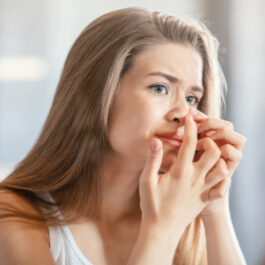 O femeie tânără cu o expresie nemulțumită își presează acneea de pe nas, în timp ce se uită în oglindă