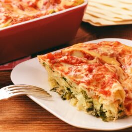Porție de Lasagna cu spanac și brânzeturi pe o farfurie cu furculiță, alături de vasul cu lasagna