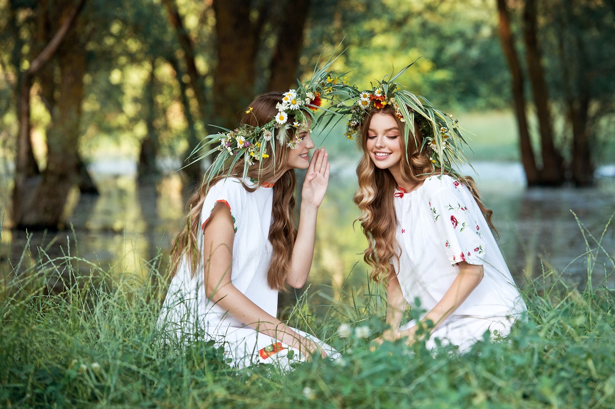 Două fete frumoase care poartă haine tradiționale românești și coronițe din flori de Sânziene