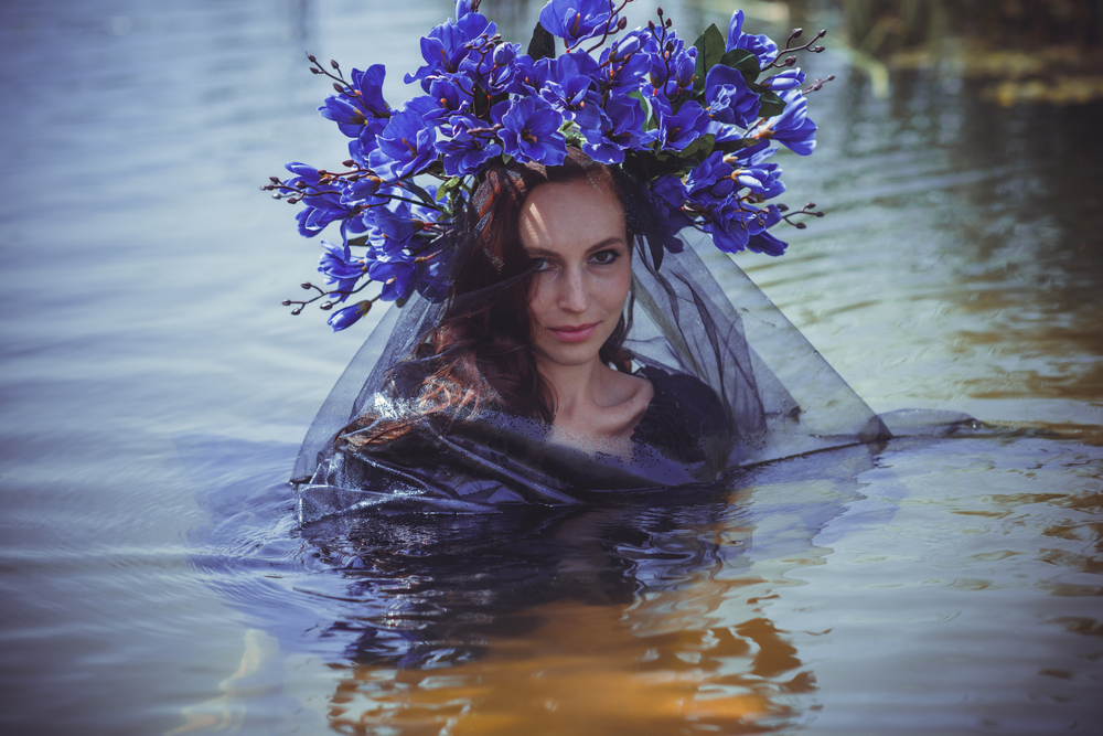 O femeie într-o rochie vaporoasă, într-un lac, cu o coroană mare din flori albastre pe cap