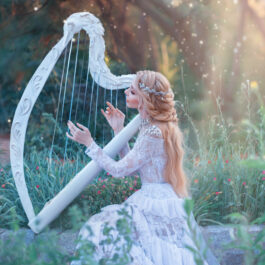 O femeie tânără, îmbrăcată într-o rochie albă cântă la harpă în pădure
