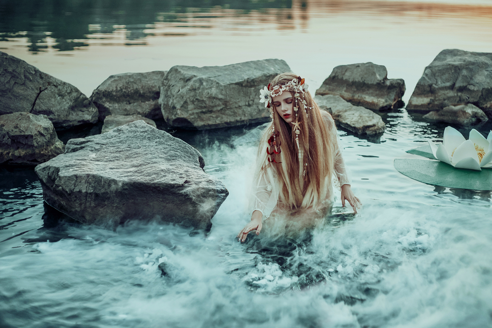 O tânără este înconjurată de crini și stânci, în apele unui lac