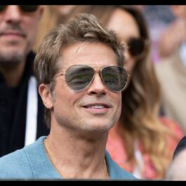 Îmbrăcat într-un tricou albastru marin și cu ochelari de soare, Brad Pitt zâmbește în timp ce se află în tribunele unui eveniment sportiv
