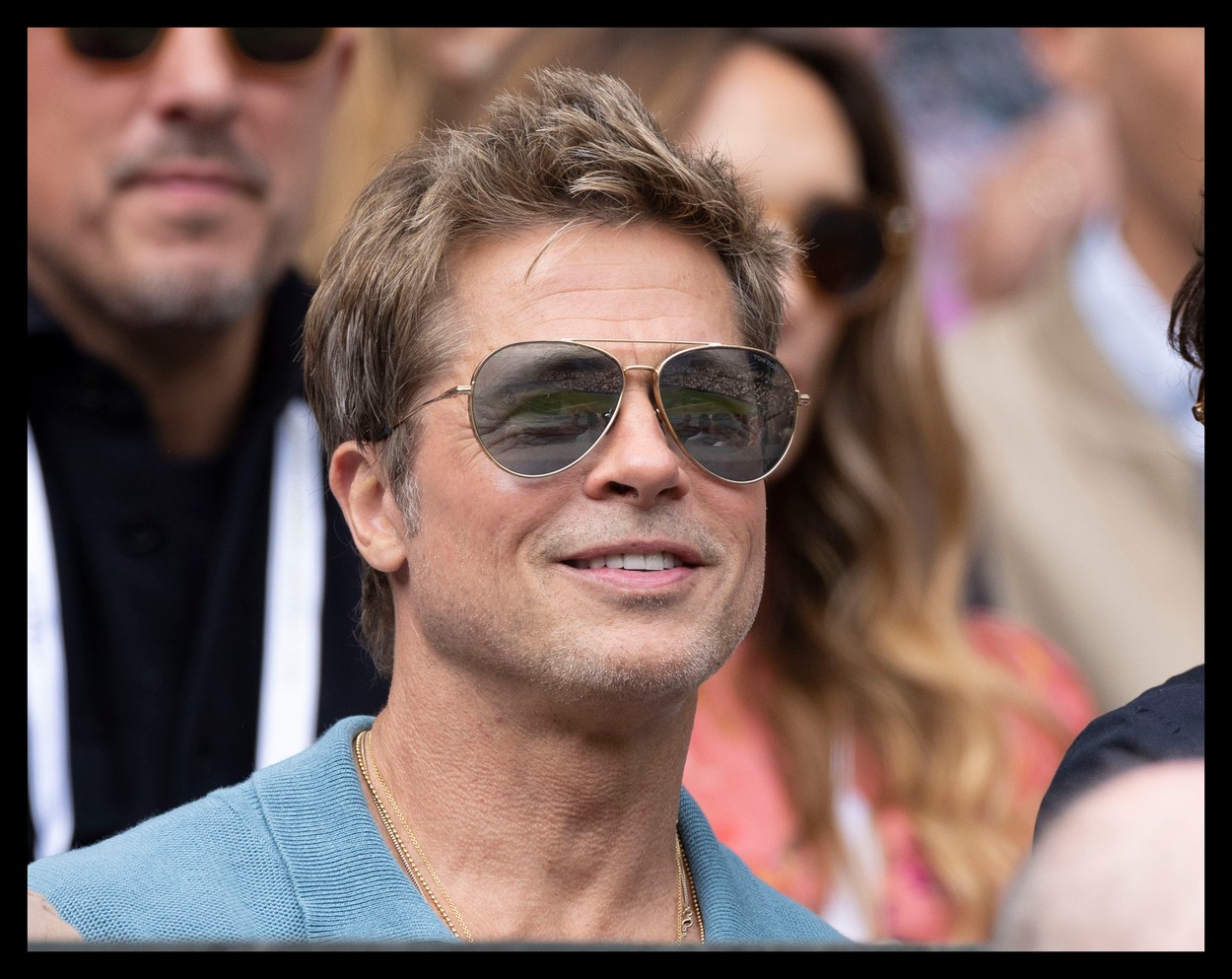 Îmbrăcat într-un tricou albastru marin și cu ochelari de soare, Brad Pitt zâmbește în timp ce se află în tribunele unui eveniment sportiv