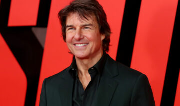 Tom Cruise, în costum negru, cu o cămașă de culoare neagră, fotografiat pe un fundal roșu