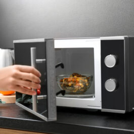O femeie folosește un cuptor cu microunde pe o masă dintr-o bucătărie, pentru a încălzi un bol cu mâncare