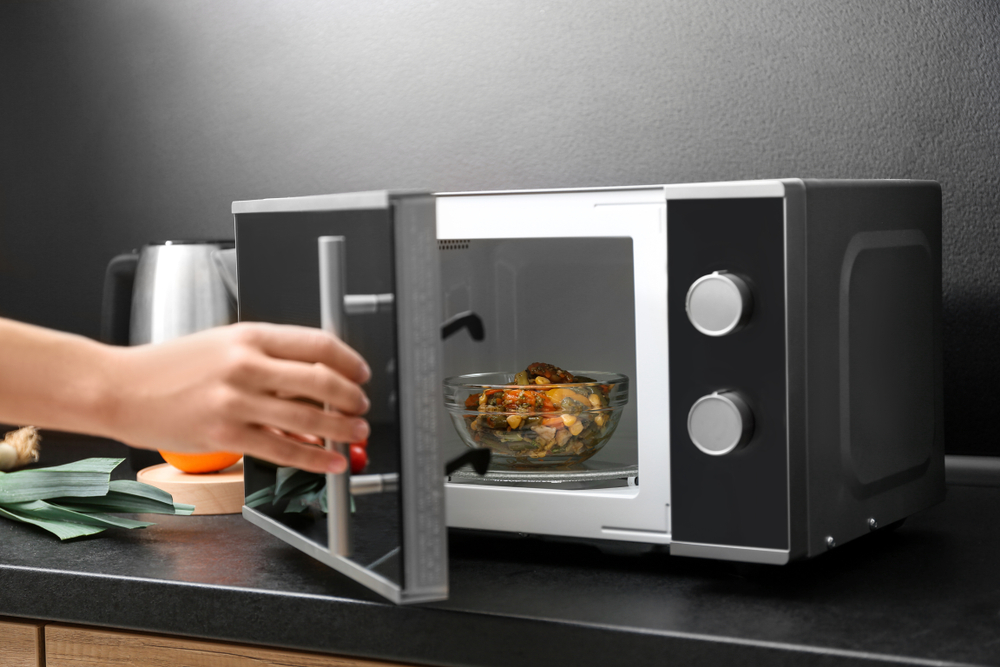 O femeie folosește un cuptor cu microunde pe o masă dintr-o bucătărie, pentru a încălzi un bol cu mâncare