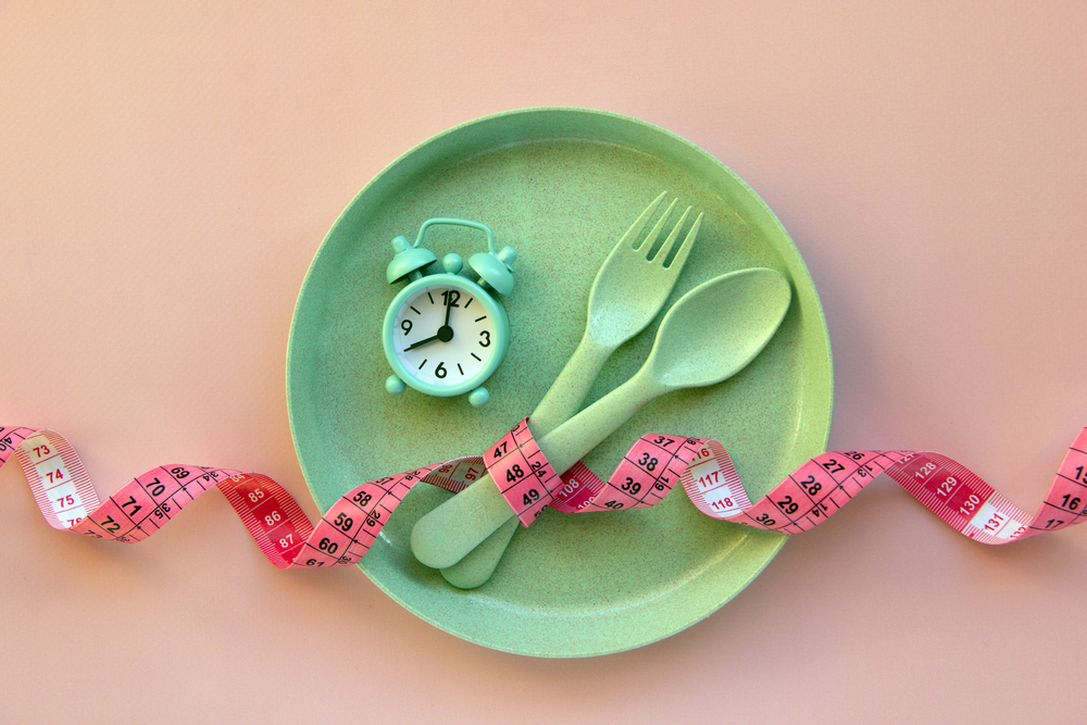 O furculiță, o lingură, un ceas și un centimetru puse pe o farfurie verde, cu un fundal roz