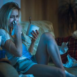 O femeie care stă în pijamale pe canapea, cu o caserola de înghețată în mână și plânge
