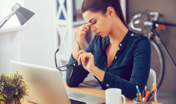 O femeie tânără obosită și stresată stă la un birou, își masează nasul în timp ce își ține ochii închiși