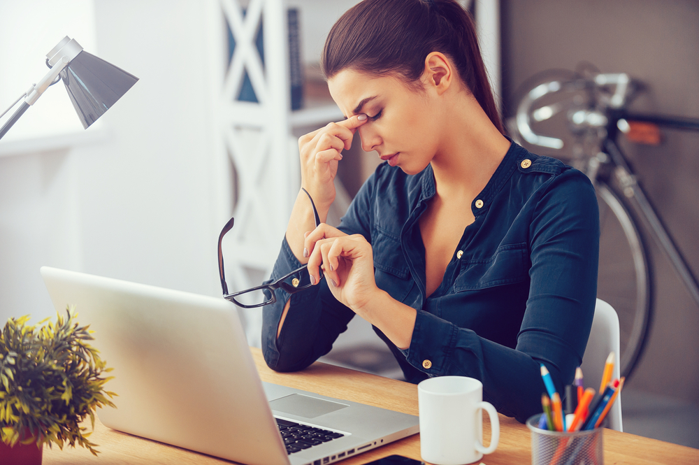 O femeie tânără obosită și stresată stă la un birou, își masează nasul în timp ce își ține ochii închiși