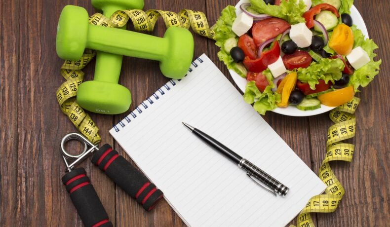 O masă de lemn pe care se află un bol cu o salată, un carnețel de notițe și câteva greutăți pentru a ilustra cum poți pierde în greutate după vârsta de 40 de ani
