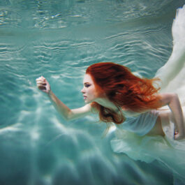 Fată frumoasă cu părul roșu înoată pe sub apă într-o rochie albă, de voal