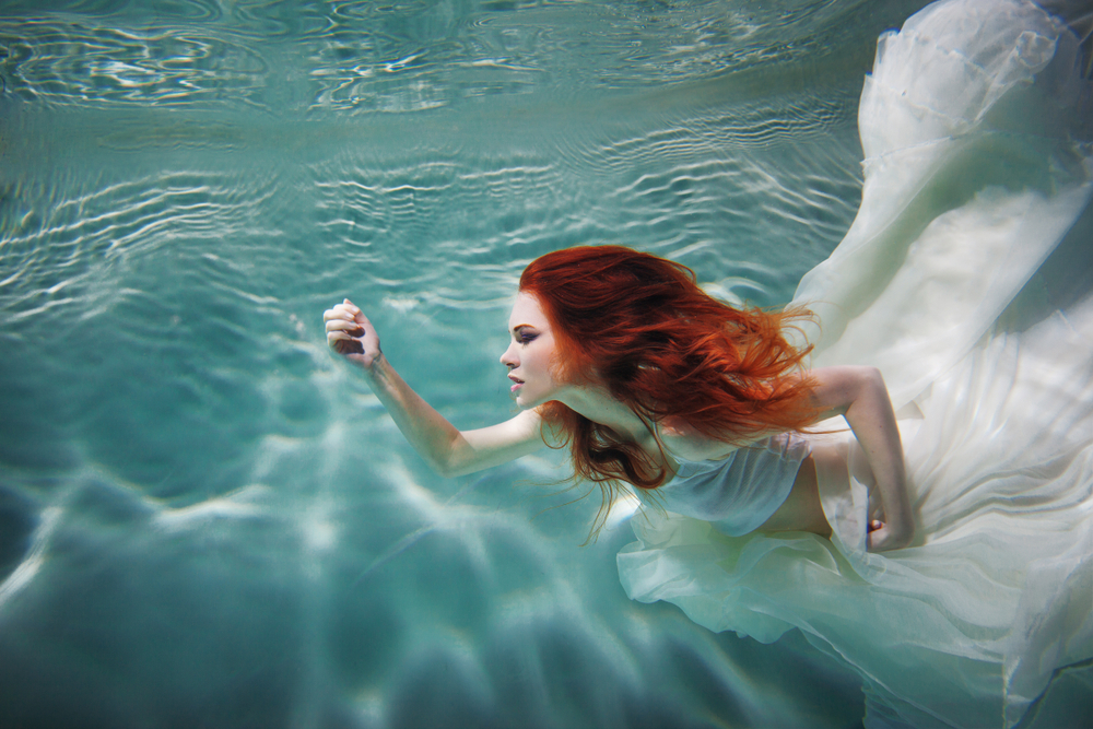 Fată frumoasă cu părul roșu înoată pe sub apă într-o rochie albă, de voal