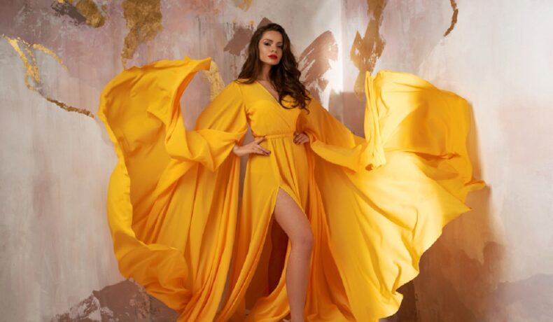 Îmbrăcată într-o rochie galbenă, elegantă și sexy, o brunetă pozează într-o cameră cu pereți golden