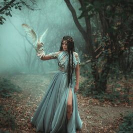 O tânără brunetă cu părul lung, îmbrăcată într-o rochie bleu de mătase care se plimbă printr-o pădure cu o bufniță albă pe mâna dreaptă