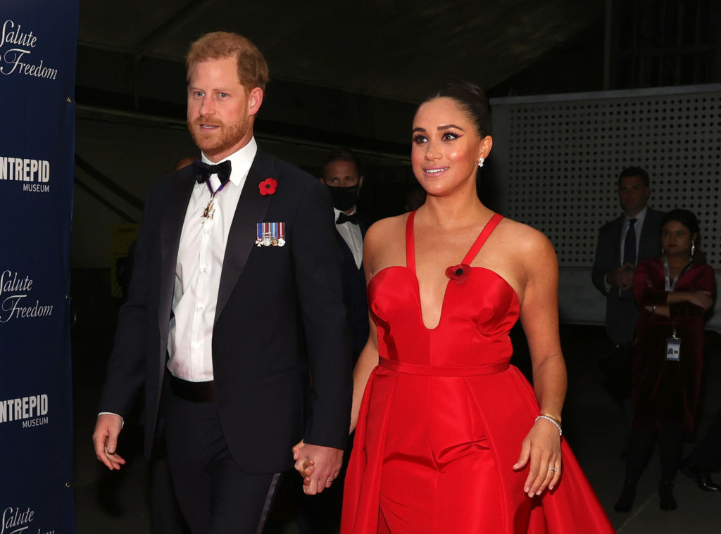 Prințul Harry este îmbrăcat la costum negru, cămașă albă și papion, iar Meghan Markle poartă o rochie elegantă, roșie, cu un decolteu adânc. Ei se țin de mână în timp ce se aflau la un eveniment organizat în 2021, în New York City
