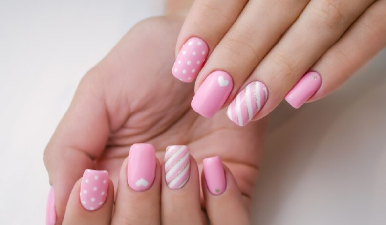 Mâinile unei tinere femei care are unghiile făcute cu gel, iar modelele sunt cu roz și alb și au și inimi