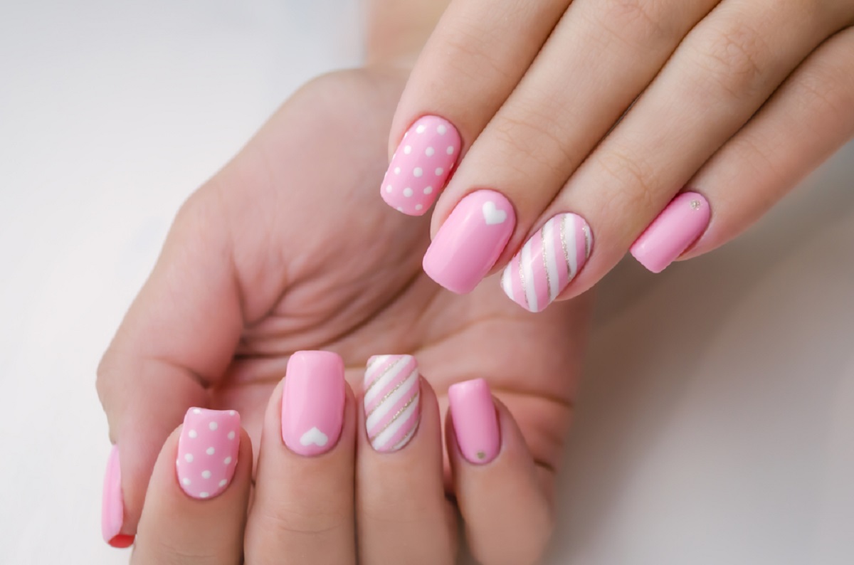 Mâinile unei tinere femei care are unghiile făcute cu gel, iar modelele sunt cu roz și alb și au și inimi