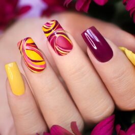 Mâna unei femei pe câteva flori roz și galbene, iar unghiile sale au nuanțe în ton cu florile
