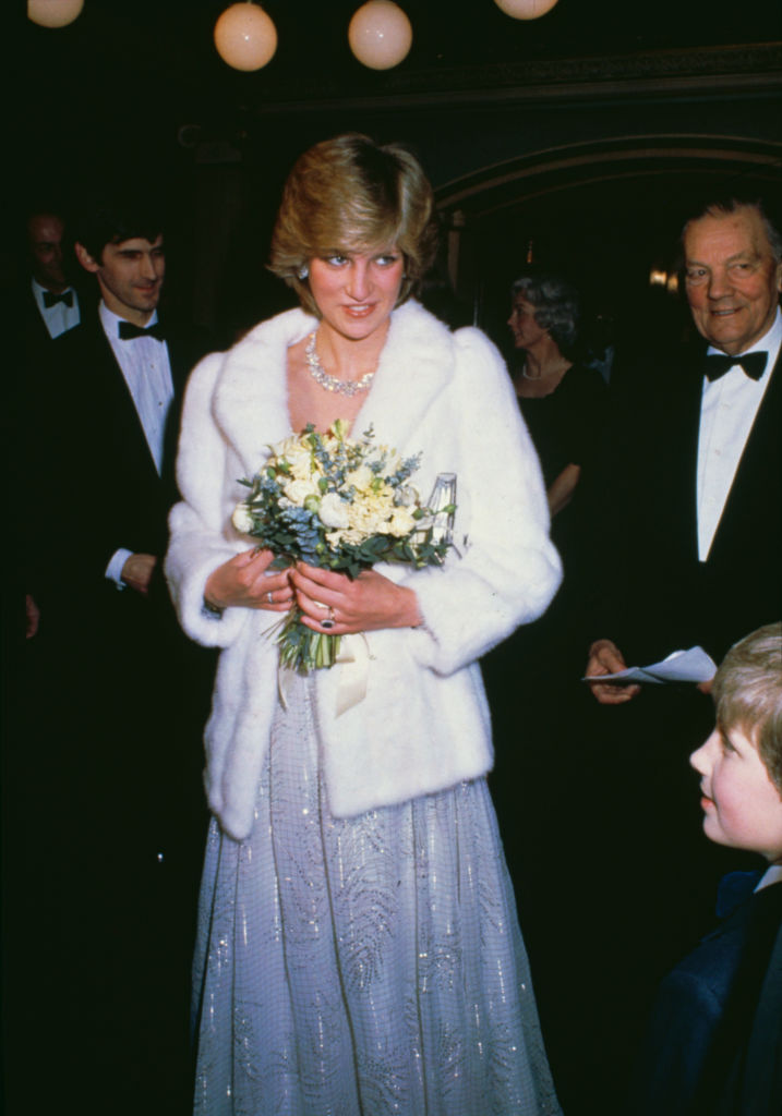 Prințesa Diana este îmbrăcată cu o rochie albă, elegantă și lungă, peste care are o haină de blană, iar în mâini ține un buchet de flori și i se vede inelul de logodnă