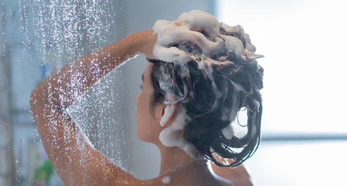 O tânără brunetă care își spală părul la duș, iar fotografia a fost făcută în timp ce ea stă cu spatele