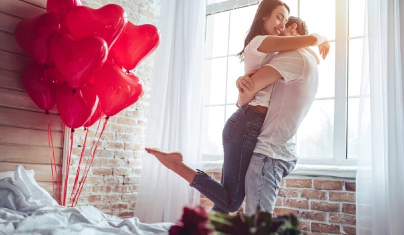 Doi tineri îndrăgostiți care se îmbrățișează în dormitor și zâmbesc de fericire, iar pe pat sunt mai multe baloane roșii, care au formă de inimă și un buchet de trandafiri roșii