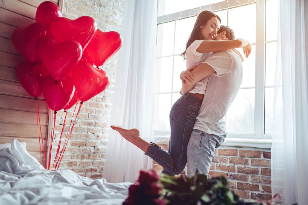 Doi tineri îndrăgostiți care se îmbrățișează în dormitor și zâmbesc de fericire, iar pe pat sunt mai multe baloane roșii, care au formă de inimă și un buchet de trandafiri roșii