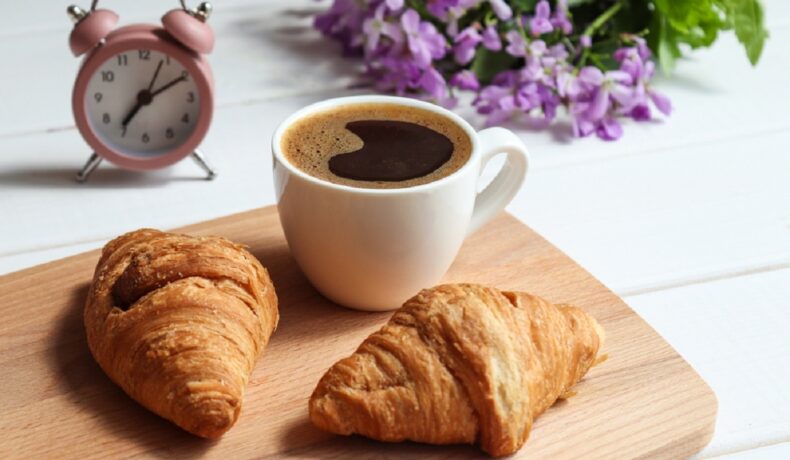 O masă pe care se află un ceas, alături de un tocător pe care sunt puse două cornuri și o ceașcă de cafea neagră pentru a ilustra în ce moment al zilei este recomandat să bei cafea pentru a pierde în greutate