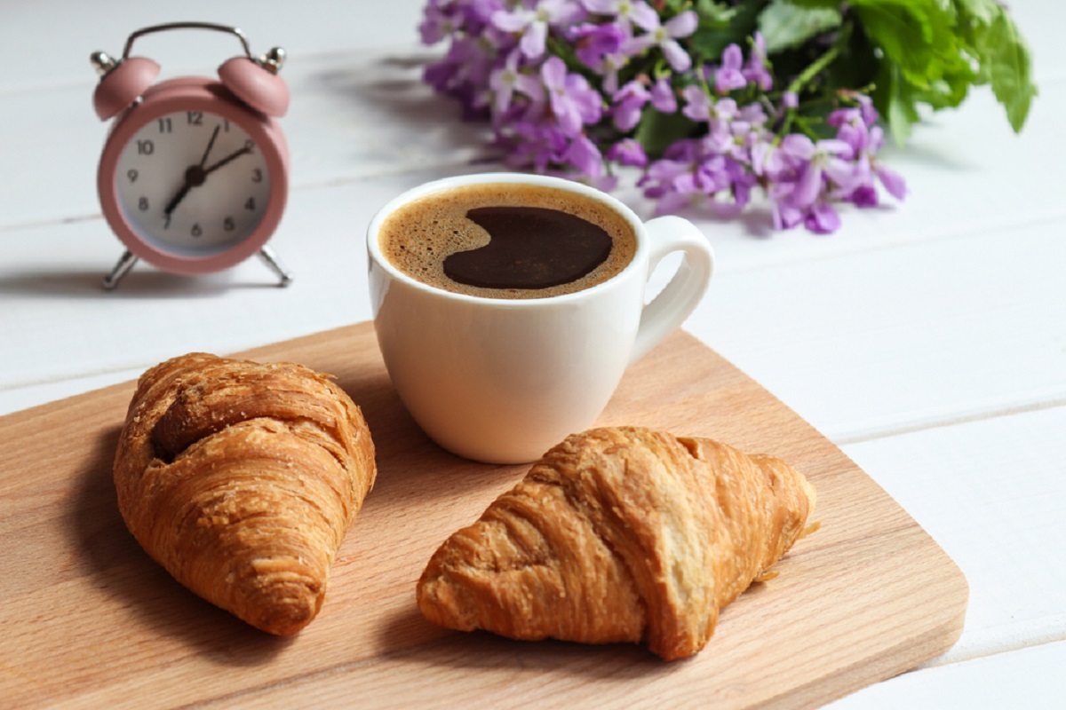 O masă pe care se află un ceas, alături de un tocător pe care sunt puse două cornuri și o ceașcă de cafea neagră pentru a ilustra în ce moment al zilei este recomandat să bei cafea pentru a pierde în greutate