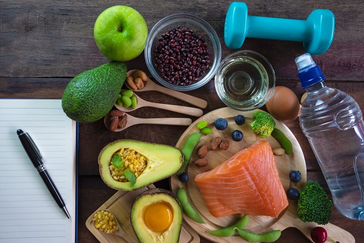 O masă pe care se află un carnet de notițe, avocado și alte fructe care te ajută să slăbești, alături de un castron cu somon și o sticlă de apă