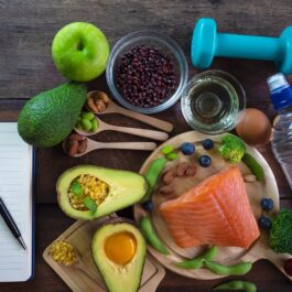 O masă pe care se află un carnet de notițe, avocado și alte fructe care te ajută să slăbești, alături de un castron cu somon și o sticlă de apă