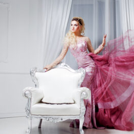 O femeie frumoasă, într-o rochie lungă, elegantă, roz, lângă o canapea albă, luxoasă