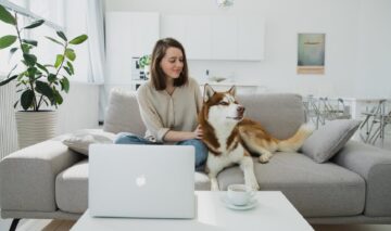 Un living spa ios în care se află o femeie cu un câine care stau împreună pe o canapea și privesc un ecran de laptop